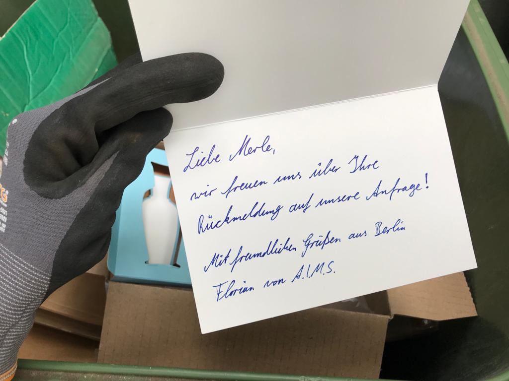 Karte mit dem Text: Liebe Merle, wir freuen uns über Ihre Rückmeldung auf unsere Anfrage! Mit freundlichen Grüßen aus Berlin Florian von A.I.M.S Im Hintergrund sieht man Papiermüll, u.a. die Verpackung eines Parfums.